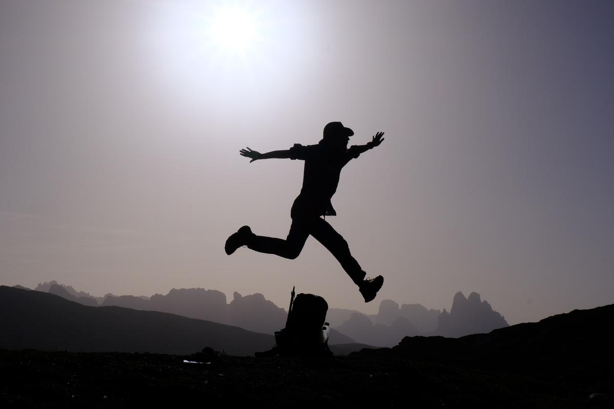 Me in mid air as I hurdle over my backpack with several peaks of the Dolomites arrayed in the background, Italy